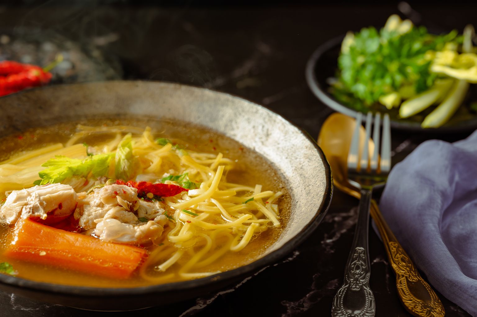 A bowl of noodle soup with fresh greens and a side dish of herbs, brass cutlery beside it — a metaphor for the daily details handled by an F&B operator.